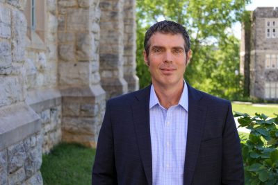 portrait of man in shirt and blazer outside next to stone building