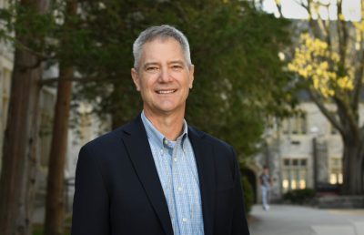 Pitts smiles outside with trees and stone buildings in background