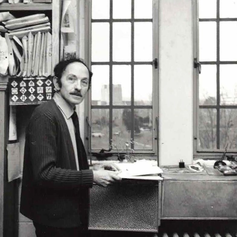 black and white photo of Good in his office by a window with Burruss Hall across the Drillfield in the background.