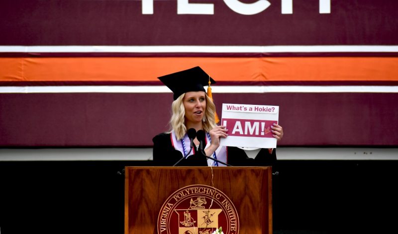 woman in graduation attire stands at podium and holds a sign that reads "what is a Hokie? I am!"